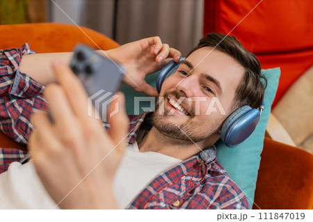 Man in wireless headphones listening energetic dancing music on smartphone relaxing lying on sofa 111847019