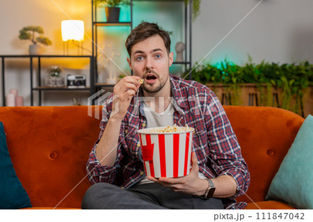 Happy man eating popcorn watching home TV shouting celebrating success winning goal game at home Happy man eating popcorn watching home TV shouting celebrating success winning goal game at home 111847042