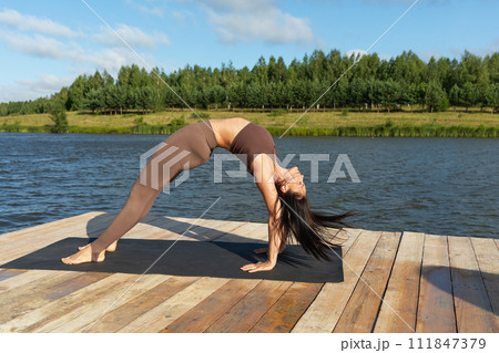 Attractive woman performing Setu Bandhasana exercise, bridge pose, working out in leggings and a short top on the shore of a lake on a sunny warm morning 111847379