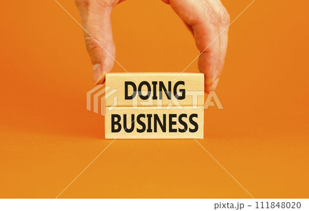 Doing business symbol. Concept words Doing business on beautiful wooden block. Beautiful orange table orange background. Businessman hand. Business, motivational Doing business concept. Copy space. Doing business symbol. Concept words Doing business on beautiful wooden block. Beautiful orange table orange background. Businessman hand. Business, motivational Doing business concept. Copy space. 111848020