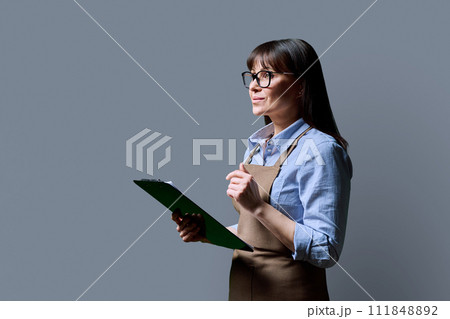 Business woman owner, worker in an apron with clipboard on gray background Business woman owner, worker in an apron with clipboard on gray background 111848892