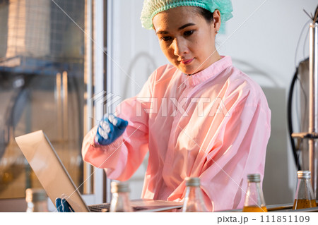 Quality manager a woman meticulously inspects beverage bottles on a factory conveyor belt. Using a laptop she ensures stringent liquid quality control. 111851109
