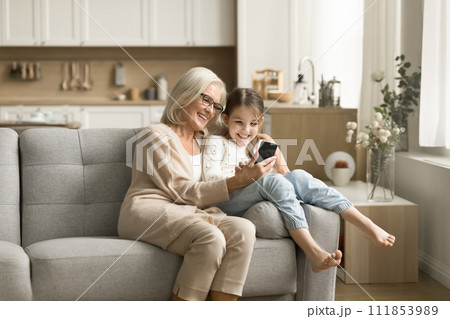 Happy granny cuddling sweet little tween grandkid girl with smartphone 111853989
