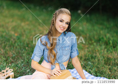Pretty caucasian young woman with long hair is sitting on a blanket in nature on a sunny summer day. Picnic, rest, relax in the fresh air. 111853990