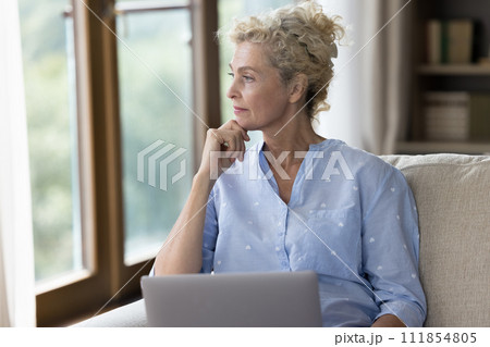 Serious older woman looking away sit on sofa with laptop 111854805