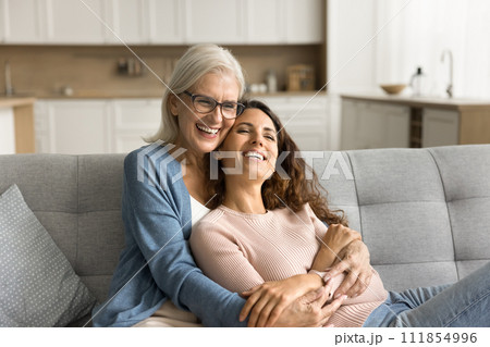 Joyful mature mother and child woman sitting together on sofa Joyful mature mother and child woman sitting together on sofa 111854996
