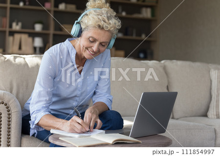 Mature woman in headphones make notes in copybook studying online Mature woman in headphones make notes in copybook studying online 111854997