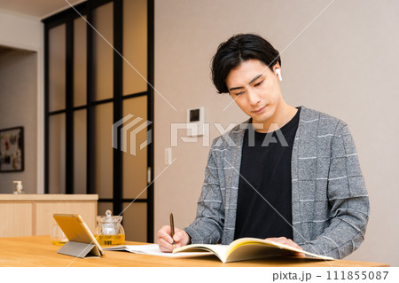 リビングで資格取得やリスキリングのためにノートパソコンを広げイヤフォンをつけ勉強をする30代の男性 リビングで資格取得やリスキリングのためにノートパソコンを広げイヤフォンをつけ勉強をする30代の男性 111855087