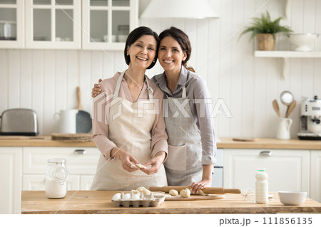 Older mother and adult daughter hugging, posing together in kitchen 111856135