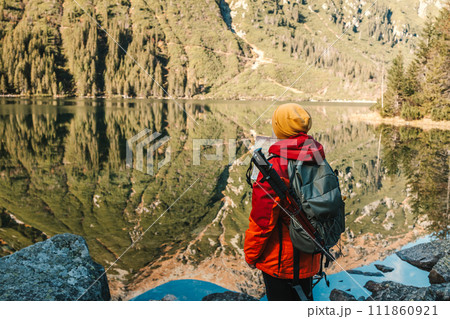 Back view of senior 50s woman in red jacket standing on top of mountain and looking at Morskie oko lake. Female with backpack on mountain edge and enjoying the beautiful view in Poland, Tatry Back view of senior 50s woman in red jacket standing on top of mountain and looking at Morskie oko lake. Female with backpack on mountain edge and enjoying the beautiful view in Poland, Tatry 111860921