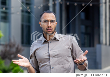 Close-up portrait of a young hispanic man wearing glasses standing outside an office building, looking worriedly at the camera and spreading his arms. Close-up portrait of a young hispanic man wearing glasses standing outside an office building, looking worriedly at the camera and spreading his arms. 111864461