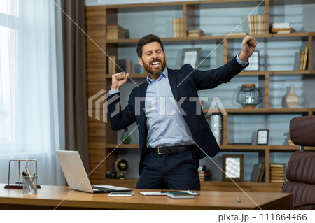A happy young man in a business suit standing in the office at the desk with a laptop and dancing with his hands up, rejoicing at the success and result, resting during a break. 111864466
