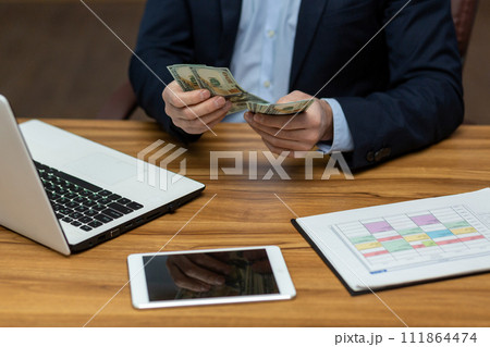 Photo concept close-up of the hand of a young man in a business suit sitting in the office at a table with gadgets, holding and counting cash dollar bills. Photo concept close-up of the hand of a young man in a business suit sitting in the office at a table with gadgets, holding and counting cash dollar bills. 111864474