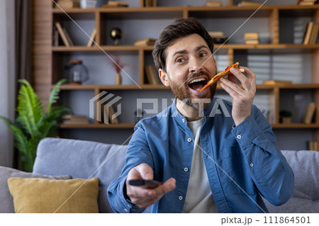 Happy man eating a slice of pizza and holding a remote control, with a content smile as he enjoys leisure time. Happy man eating a slice of pizza and holding a remote control, with a content smile as he enjoys leisure time. 111864501