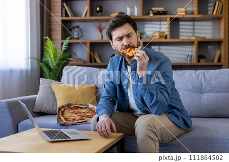 Professional male telecommuting takes a lunch break, enjoying a slice of delicious pizza while browsing on laptop. 111864502