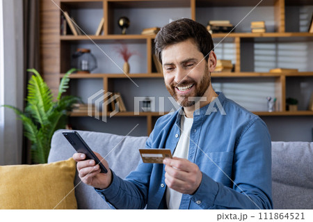 Content male in casuals holding a credit card and smartphone, purchasing online with a cheerful expression. Content male in casuals holding a credit card and smartphone, purchasing online with a cheerful expression. 111864521