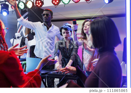 Electronic music band performing on stage in nightclub, while people partying on dancefloor. Woman dj using tracks mixer and singer giving mic to excited crowded to sing along Electronic music band performing on stage in nightclub, while people partying on dancefloor. Woman dj using tracks mixer and singer giving mic to excited crowded to sing along 111865865