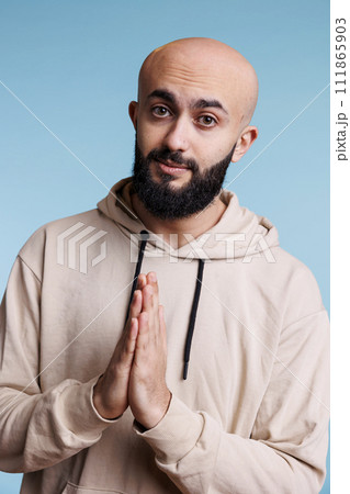 Arab man praying with hands folded in worship, expressing faith studio portrait. Young adult bald bearded person pleading, making wish and looking at camera with hopeful emotions Arab man praying with hands folded in worship, expressing faith studio portrait. Young adult bald bearded person pleading, making wish and looking at camera with hopeful emotions 111865903