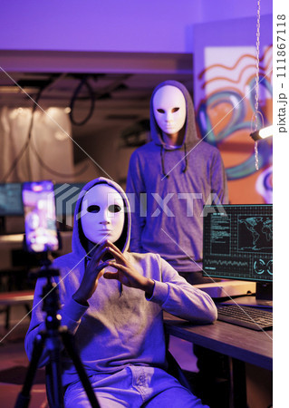 Scammers in anonymous masks streaming threat online and demand victim for payment. Hackers with hidden identity filming ransom message on mobile phone in abandoned warehouse Scammers in anonymous masks streaming threat online and demand victim for payment. Hackers with hidden identity filming ransom message on mobile phone in abandoned warehouse 111867118