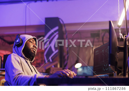 Relaxed african american criminal in hood hacking computer server while listening to music in headphones. Man hacker in earphones wearing hood planning phishing online scam 111867286