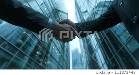 Close-up of businessmen shaking hands with a partner, against a finance and investment background featuring glass buildings. Shot from below with shallow depth of field. 111872448