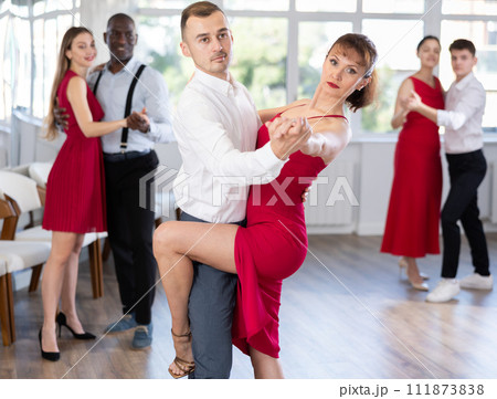 Smiling young woman wearing red dress dancing in couple rumba dance with her partner during party at dance salon Smiling young woman wearing red dress dancing in couple rumba dance with her partner during party at dance salon 111873838
