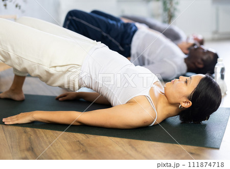 Asian woman performing asana Sarvangasana during group yoga workout Asian woman performing asana Sarvangasana during group yoga workout 111874718