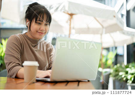 A positive Asian woman working remotely at a cafe in the city, using her laptop. A positive Asian woman working remotely at a cafe in the city, using her laptop. 111877724