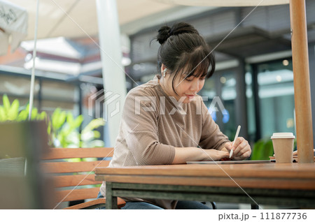 A focused Asian woman is working remotely at a cafe, wearing earbuds and using her digital tablet. 111877736