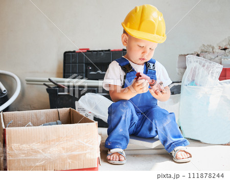 Cute little boy sitting on the floor near cardboard box with tools and materials for home renovation. Kid construction worker in safety helmet holding construction component in apartment. 111878244