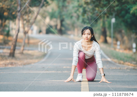 Fitness concept, A focused female runner is in the starting position on a serene, tree-flanked road, poised for a morning run. 111878674
