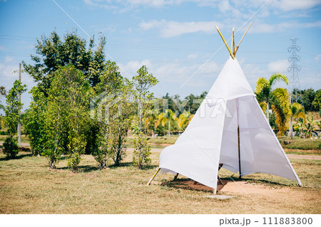 A charming summer wedding teepee stands in a green field evoking joy and celebration. The traditional wigwam adorned with dreamcatcher decorations creates a picturesque scene of family happiness. A charming summer wedding teepee stands in a green field evoking joy and celebration. The traditional wigwam adorned with dreamcatcher decorations creates a picturesque scene of family happiness. 111883800