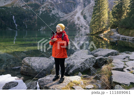 Rear shot of traveler senior woman in sportswear standing on the rocky shore and making selfie on smart phone or video. Taking photo of green hills and mountains on Morskie Oko lake. Hiking travel and Rear shot of traveler senior woman in sportswear standing on the rocky shore and making selfie on smart phone or video. Taking photo of green hills and mountains on Morskie Oko lake. Hiking travel and 111889061