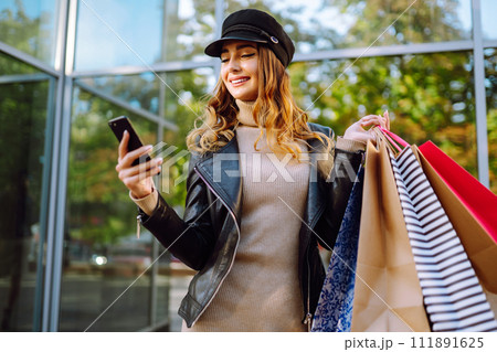 Smiling woman with shopping bags using her phone. Young woman after shopping on the city street. Smiling woman with shopping bags using her phone. Young woman after shopping on the city street. 111891625