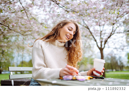 Young woman sitting at a table in blooming spring park with a notepad and laptop. Lifestyle concept. Young woman sitting at a table in blooming spring park with a notepad and laptop. Lifestyle concept. 111892332