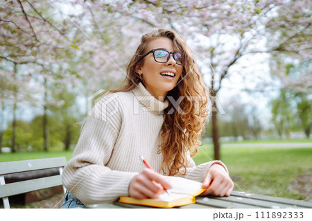 Young woman sitting at a table in blooming spring park with a notepad and laptop. Lifestyle concept. 111892333