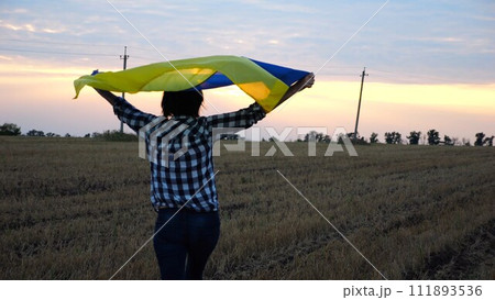 Ukrainian woman walking on barley meadow with a blue-yellow banner on shoulders at sunset. Lady going on wheat field with a flag of Ukraine as symbol of victory against rusian aggression. Slow mo 111893536