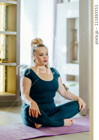 Vertical shot of full-figured mature middle age woman sitting on mat in lotus pose. Calm lady practicing techniques and yoga indoors. Vertical shot of full-figured mature middle age woman sitting on mat in lotus pose. Calm lady practicing techniques and yoga indoors. 111893551