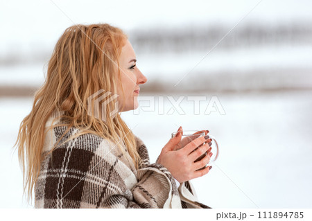 Side view portrait of red-haired pretty girl with cup in her hands outdoors. Girl enjoys snowy winter day. 111894785