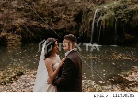 bride in white wedding dress and long veil hugs groom in a suit small pond lake river waterfall Burbun bride in white wedding dress and long veil hugs groom in a suit small pond lake river waterfall Burbun 111898685