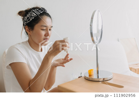 Asian Thai woman using and applying sun cream moisturizer in front of the mirror while sitting on white couch in room apartment, beauty concept. Asian Thai woman using and applying sun cream moisturizer in front of the mirror while sitting on white couch in room apartment, beauty concept. 111902956