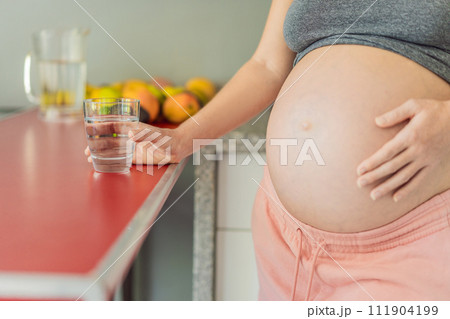 Embracing the vital benefits of water during pregnancy, a pregnant woman stands in the kitchen with a glass, highlighting hydration's crucial role in maternal well-being Embracing the vital benefits of water during pregnancy, a pregnant woman stands in the kitchen with a glass, highlighting hydration's crucial role in maternal well-being 111904199