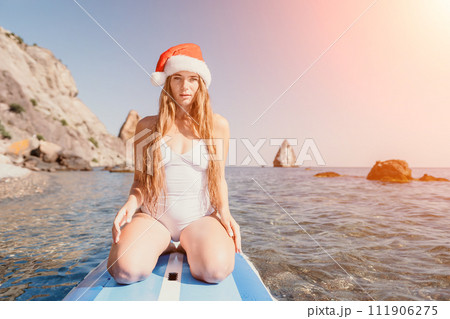 Woman sea sup. Close up portrait of happy young caucasian woman with long hair in Santa hat looking at camera and smiling. Cute woman portrait in a white bikini posing on sup board in the sea Woman sea sup. Close up portrait of happy young caucasian woman with long hair in Santa hat looking at camera and smiling. Cute woman portrait in a white bikini posing on sup board in the sea 111906275