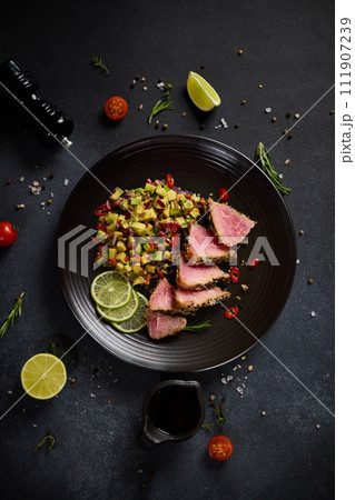 Sliced Organic grilled Tuna fillet covered with sesame seeds and salsa on black ceramic plate 111907239