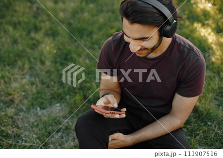 Young boy listening to music while sitting on a grass. Man using a smartphone. Man wearing a bordo shirt and jeans. 111907516