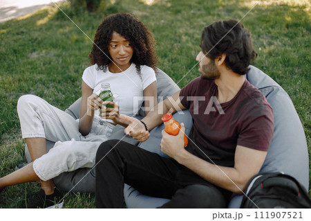 Friends hanging in the green park, relaxing, sitting on the bean bag chairs on grass. Boy and girl holding a bottles of juice. African girl wearing white clothes, boy wearing brown t-shirt and black 111907521