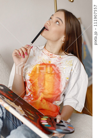 Young female artist posing holding brushes and painting in hands. Woman wearing white t-shirt and blue jeans. Brunette girl sitting in a chair in room with white wall. 111907557