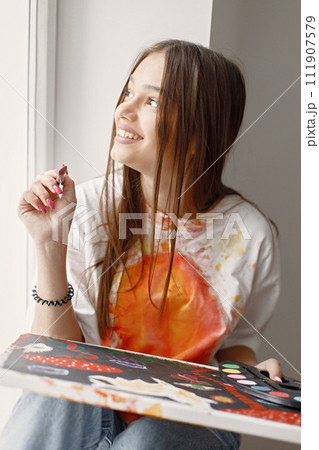 Young female artist posing holding brushes and painting in hands. Woman wearing white t-shirt and blue jeans. Brunette girl sitting on a windowsill in room with white wall. 111907579