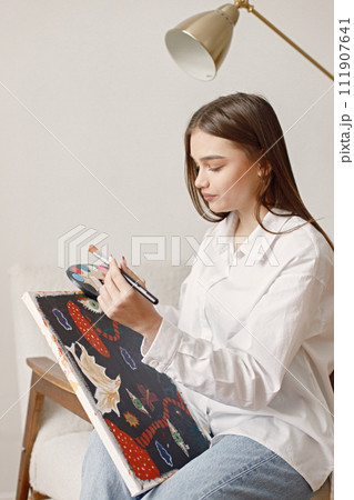 Satisfied female artist posing holding new picture and paint palette in hands. Woman wearing jeans and white shirt. Brunette girl sitting on a chair in room with white wall. Satisfied female artist posing holding new picture and paint palette in hands. Woman wearing jeans and white shirt. Brunette girl sitting on a chair in room with white wall. 111907641