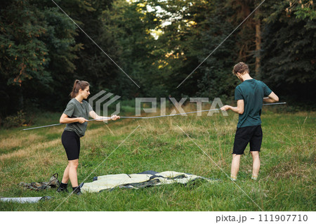 Man and woman with backpacks on a grass in forest and setting up a tent. Couple with a tent in forest at summer during their trip. Man and woman wearing green t-shirts. 111907710